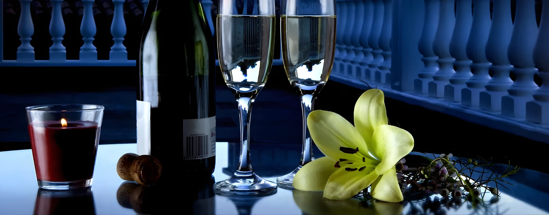 Vela, botella, copas y flor amarilla sobre mesa reflejante.