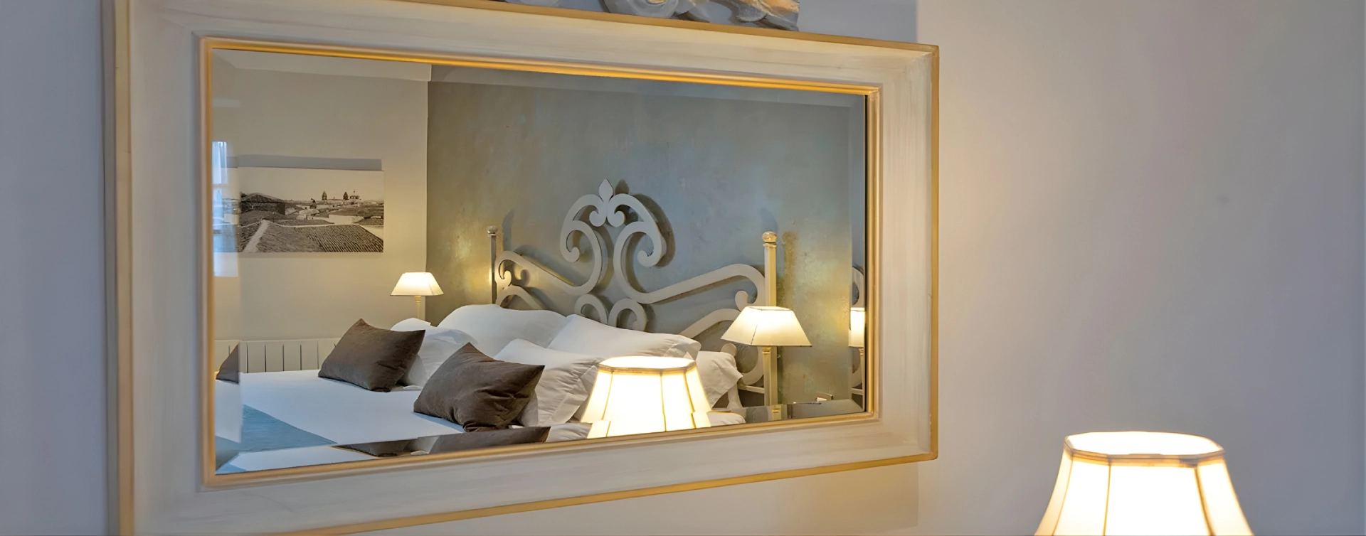 Espejo reflejando una cama con l&aacute;mparas y cojines en una habitaci&oacute;n elegante.