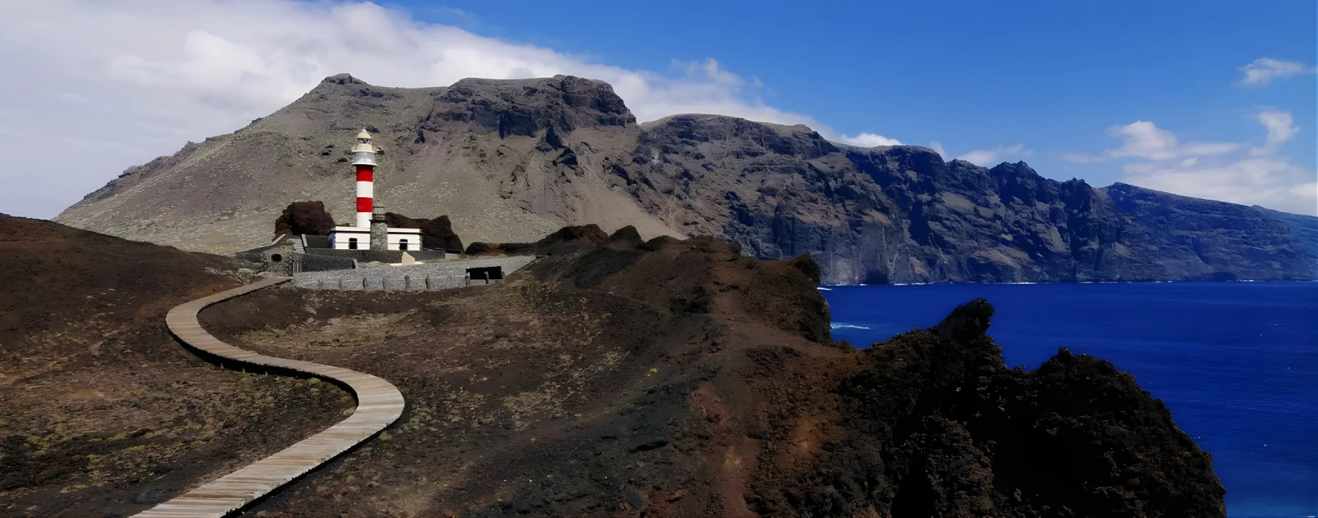 Camino de madera hacia un faro en una costa rocosa con acantilados.