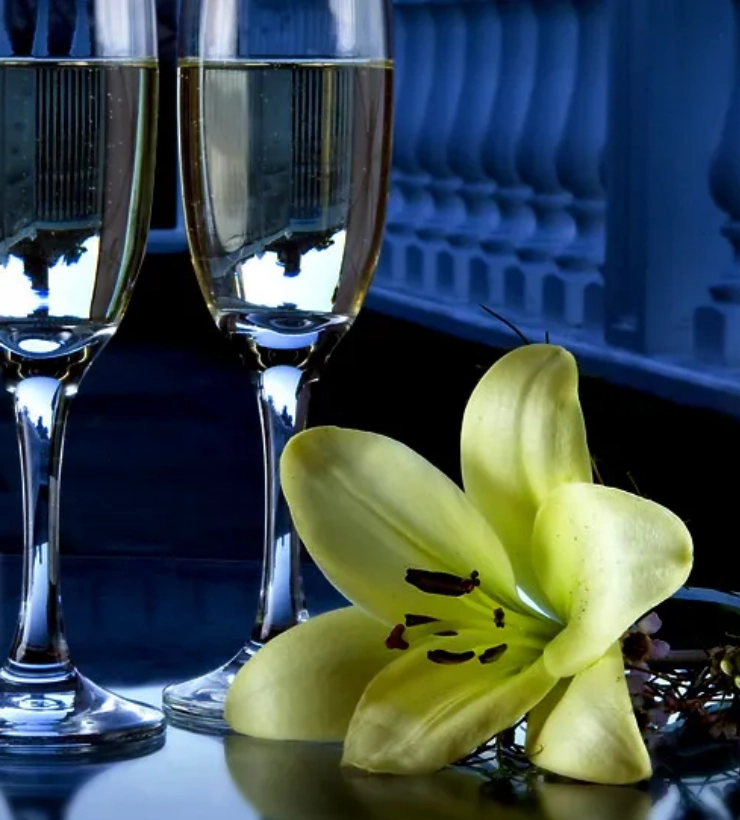 Dos copas de champ&aacute;n junto a un lirio amarillo sobre una mesa oscura.