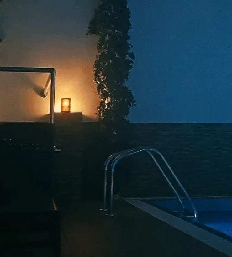 Piscina iluminada tenuemente con una vela junto a la pared y plantas decorativas.