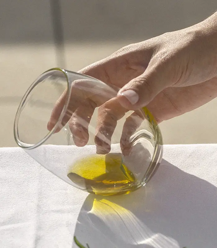 Mano inclinando un vaso de vidrio con l&iacute;quido dorado sobre un mantel blanco.
