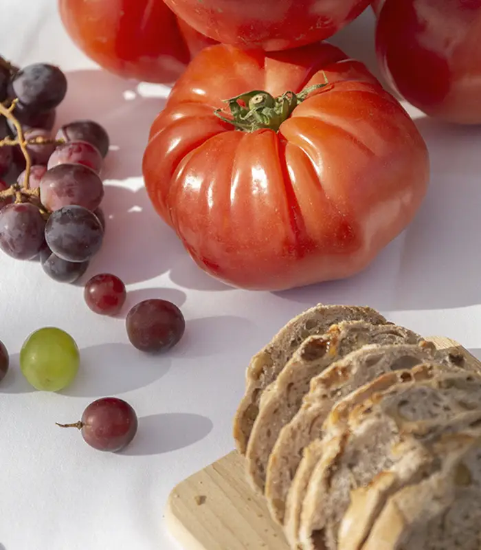 Tomates, uvas y rebanadas de pan sobre una mesa blanca.