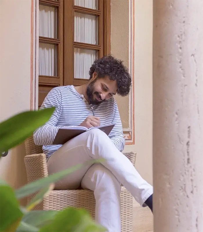 Hombre sonriente escribiendo en un cuaderno sentado en una terraza luminosa.