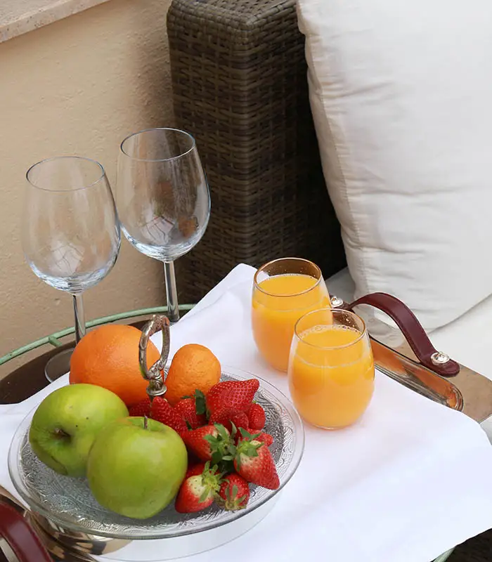 Frutas y jugos sobre una bandeja con copas vac&iacute;as y cojines de fondo.