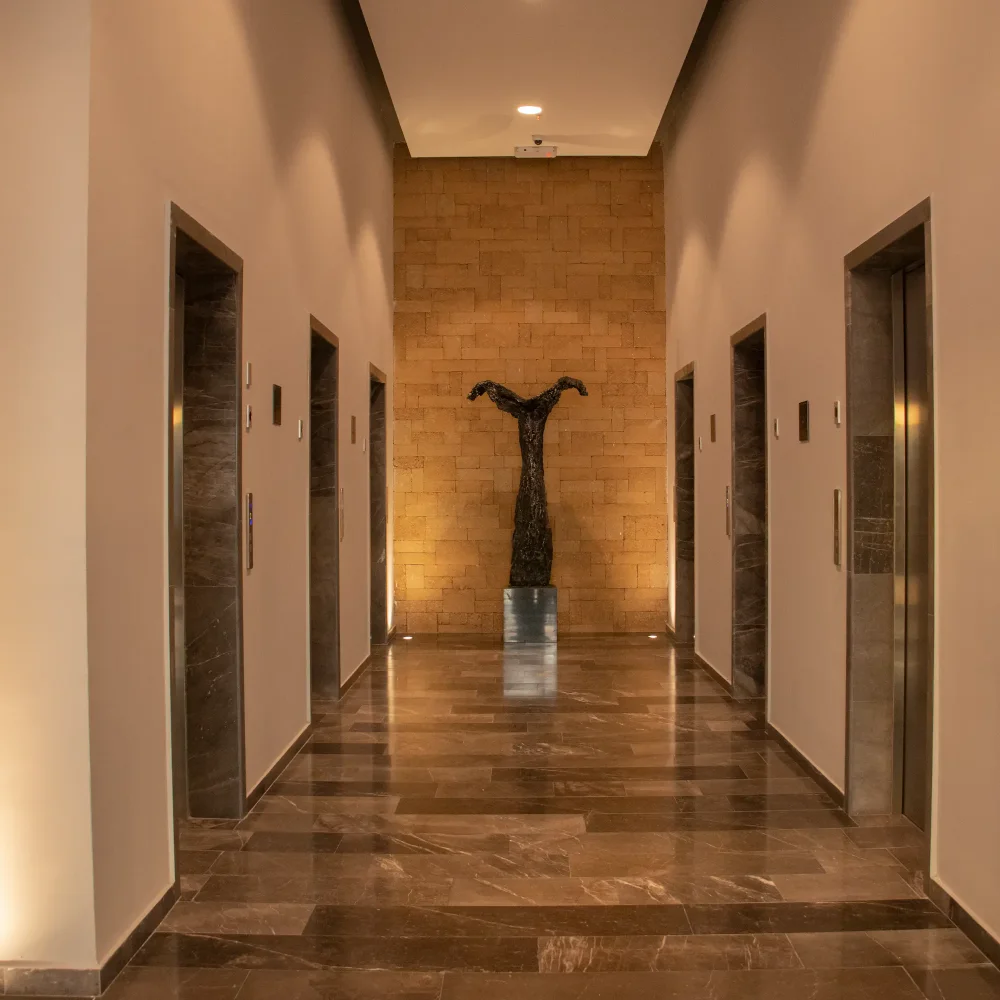 Pasillo de edificio con cuatro ascensores y una escultura moderna al fondo sobre un pedestal.