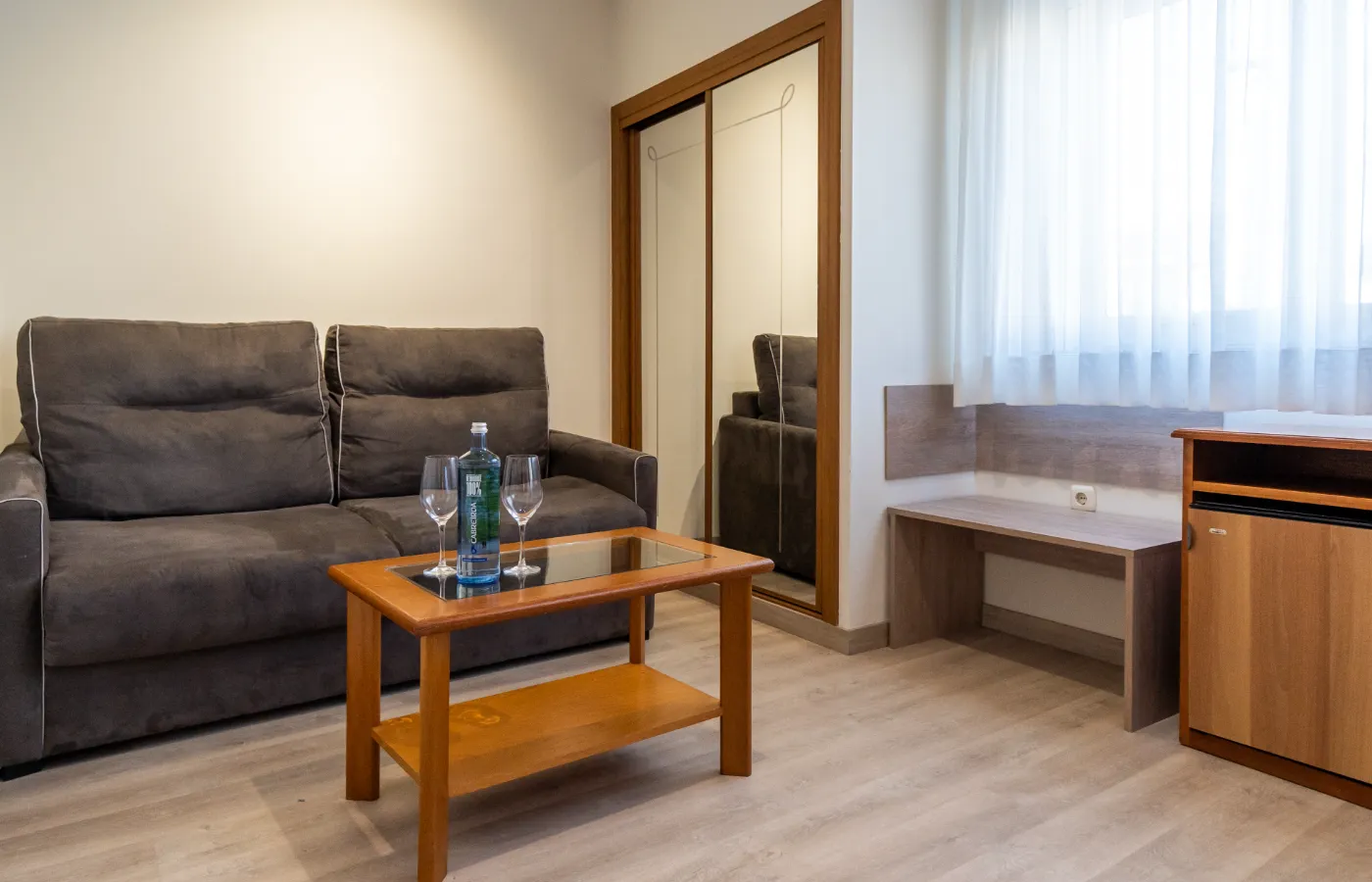 Sala con sofá gris, mesa de centro, armario con espejos y cortinas blancas.