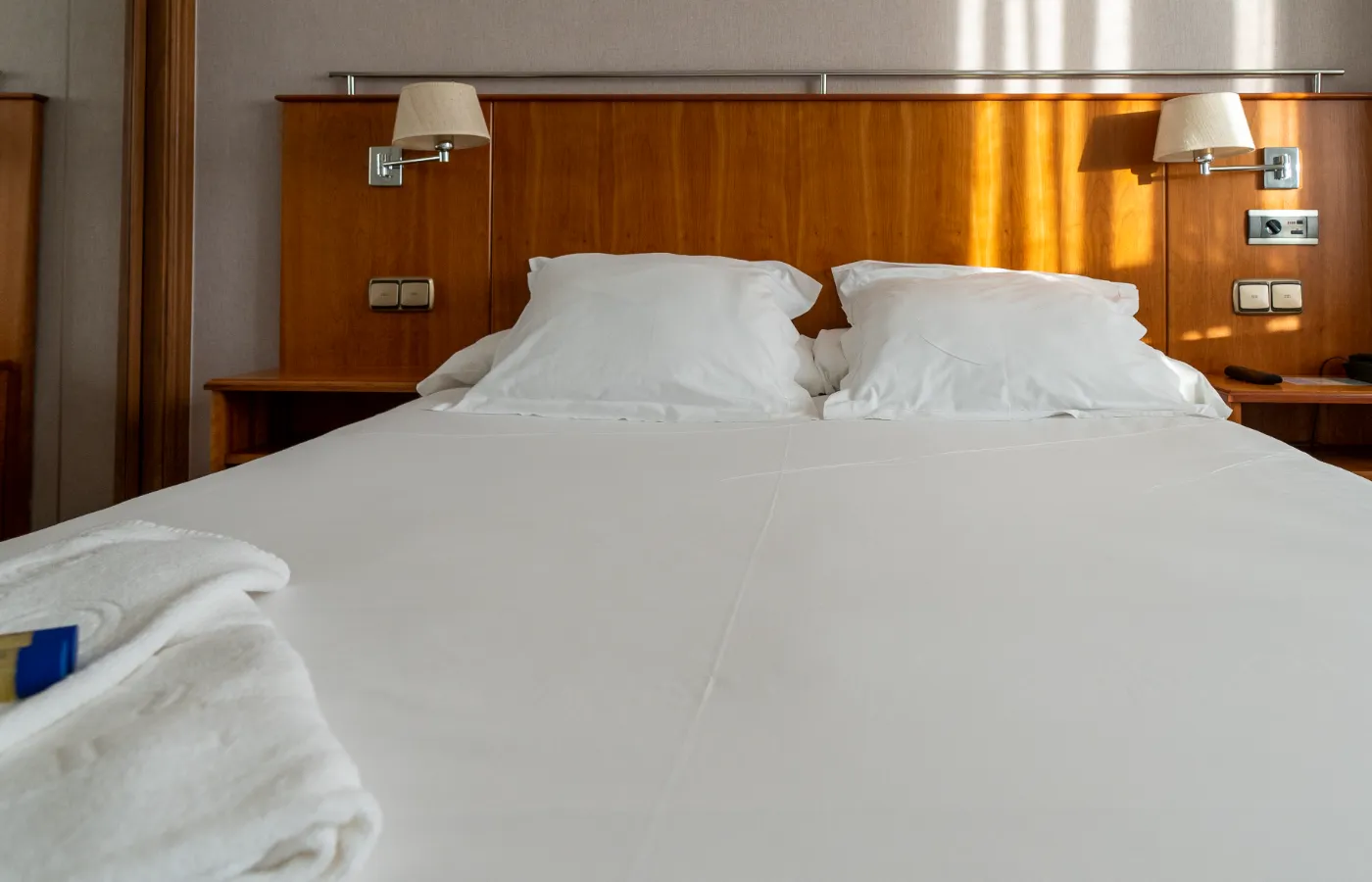 Cama matrimonial con sábanas blancas y lámparas de pared en ambos lados.
