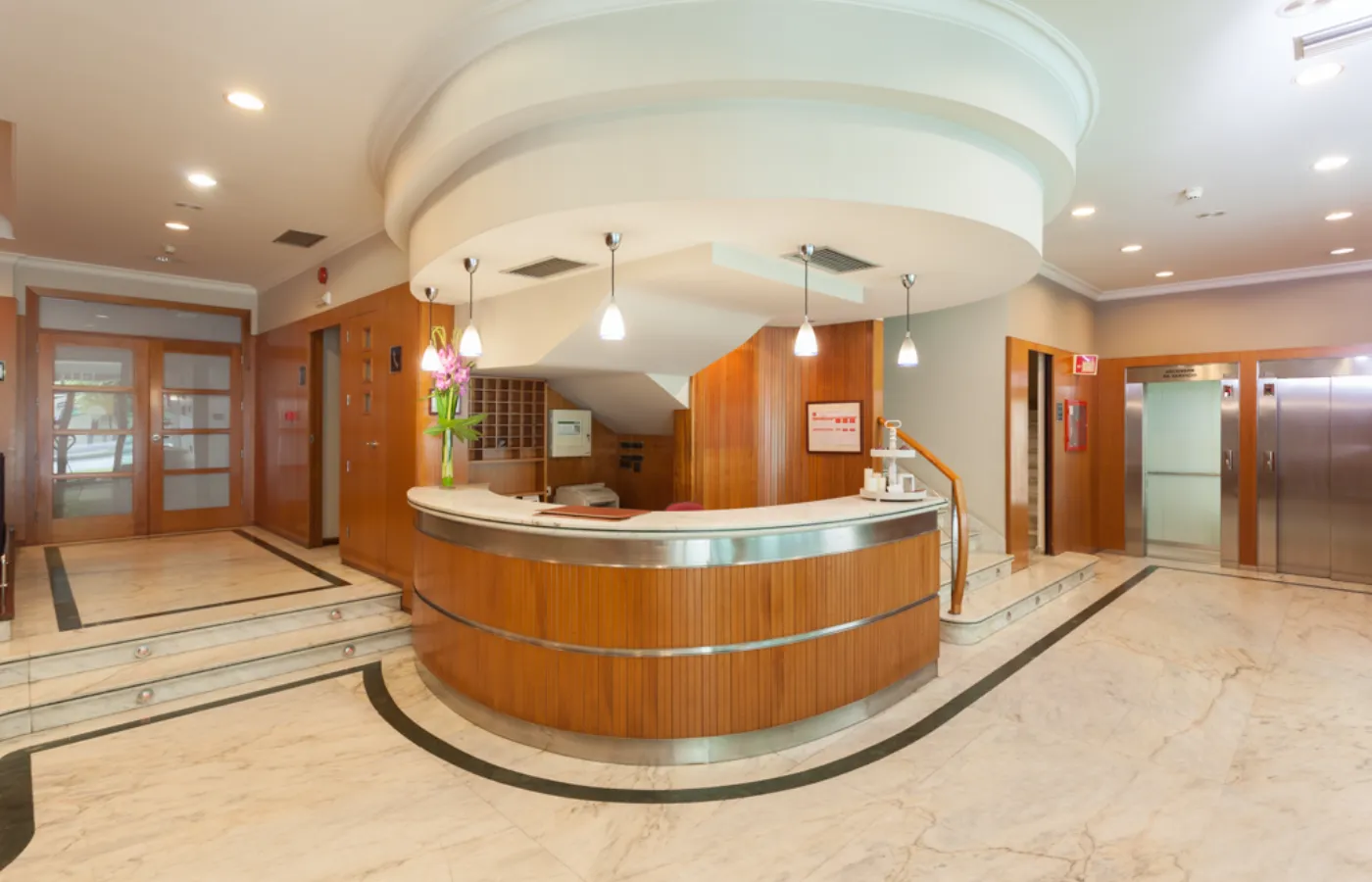 Vestíbulo con mostrador de recepción curvo, puerta y elevadores al fondo.