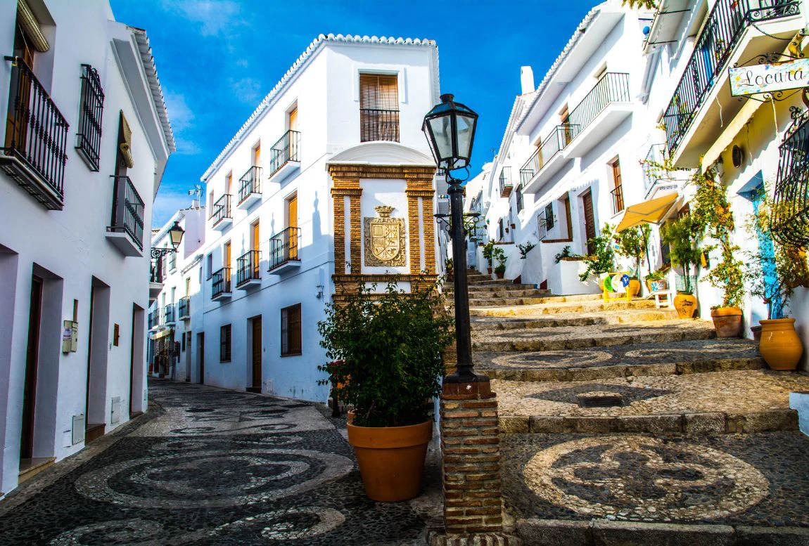 Calle empedrada con casas blancas y plantas, bajo un cielo azul.