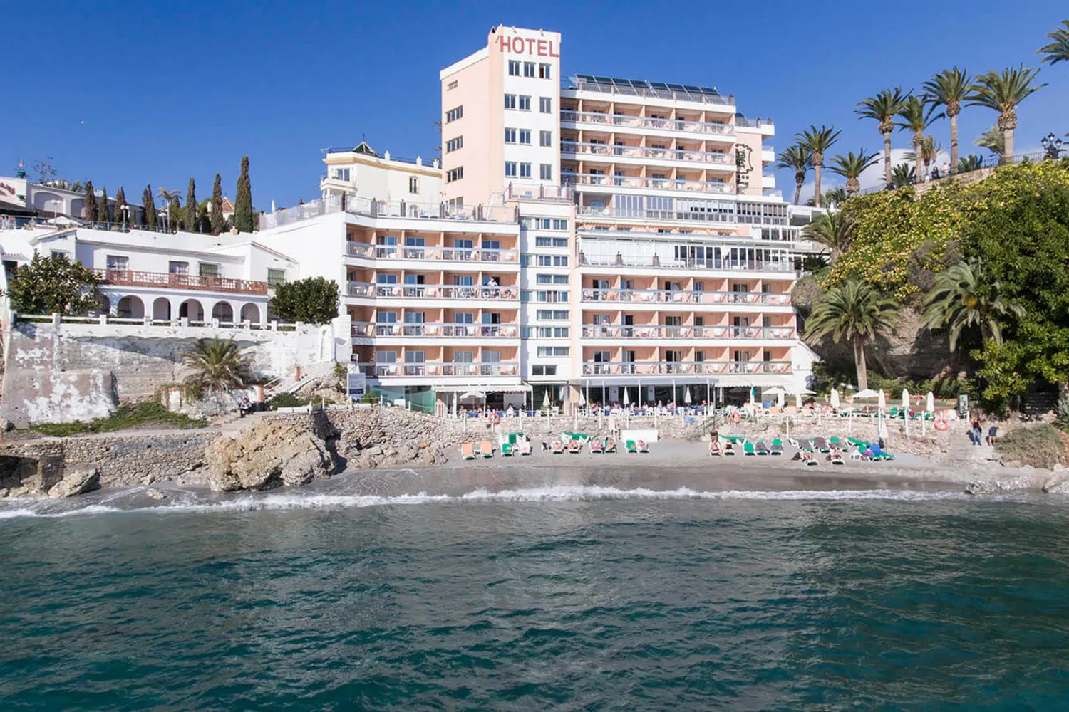 Hotel en la playa con hamacas, palmeras y cielo despejado.