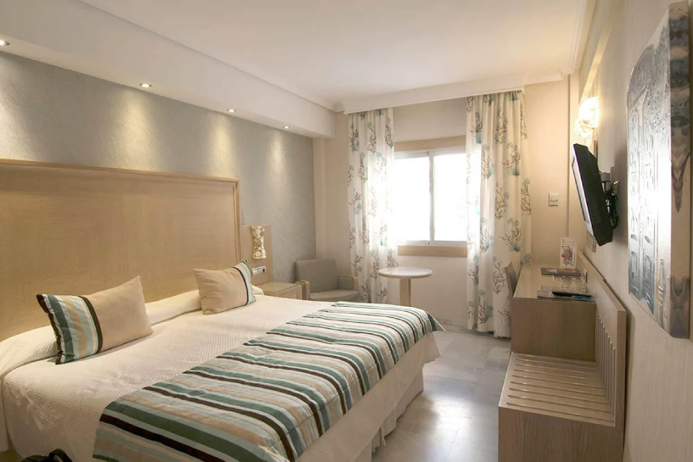 Habitaci&oacute;n de hotel con cama doble, ventana y televisi&oacute;n en la pared.