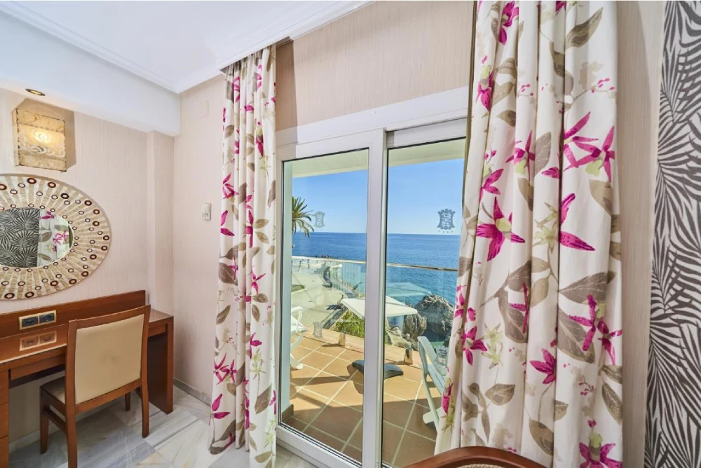 Habitaci&oacute;n con ventanal al mar, cortinas florales y escritorio con espejo.