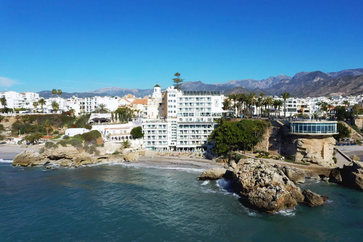 Vista costera de un hotel blanco junto al mar y monta&ntilde;as al fondo.