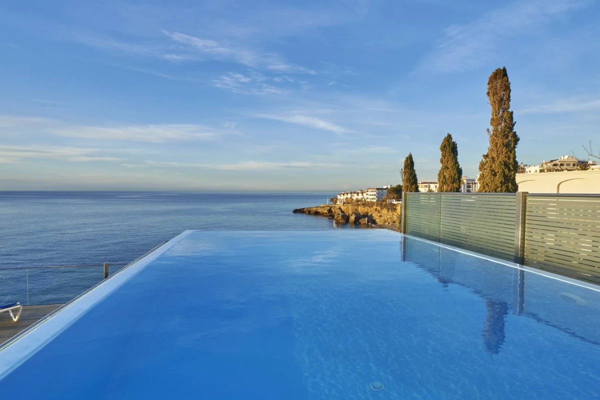 Piscina infinita con vista al mar y edificios al fondo en un d&iacute;a soleado.