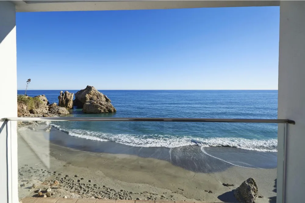 Vista al mar con playa y rocas desde un balc&oacute;n con barandilla.