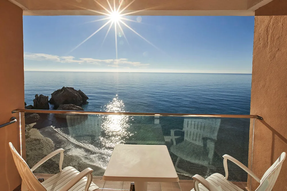 Vista al mar desde un balc&oacute;n con mesa y sillas, sol reflejado en el agua.