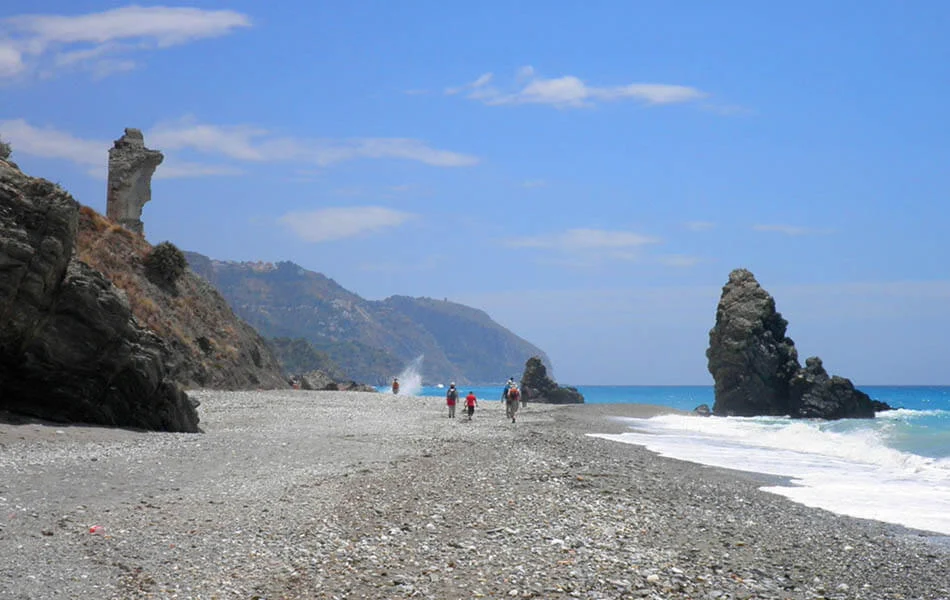 Playa rocosa con turistas, torre al fondo y mar azul bajo cielo despejado.