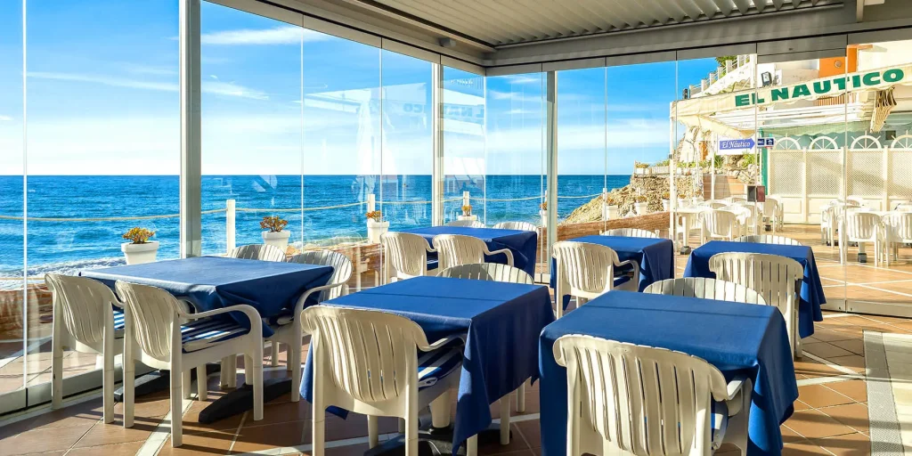 Restaurante con vista al mar, mesas con manteles azules y sillas blancas.