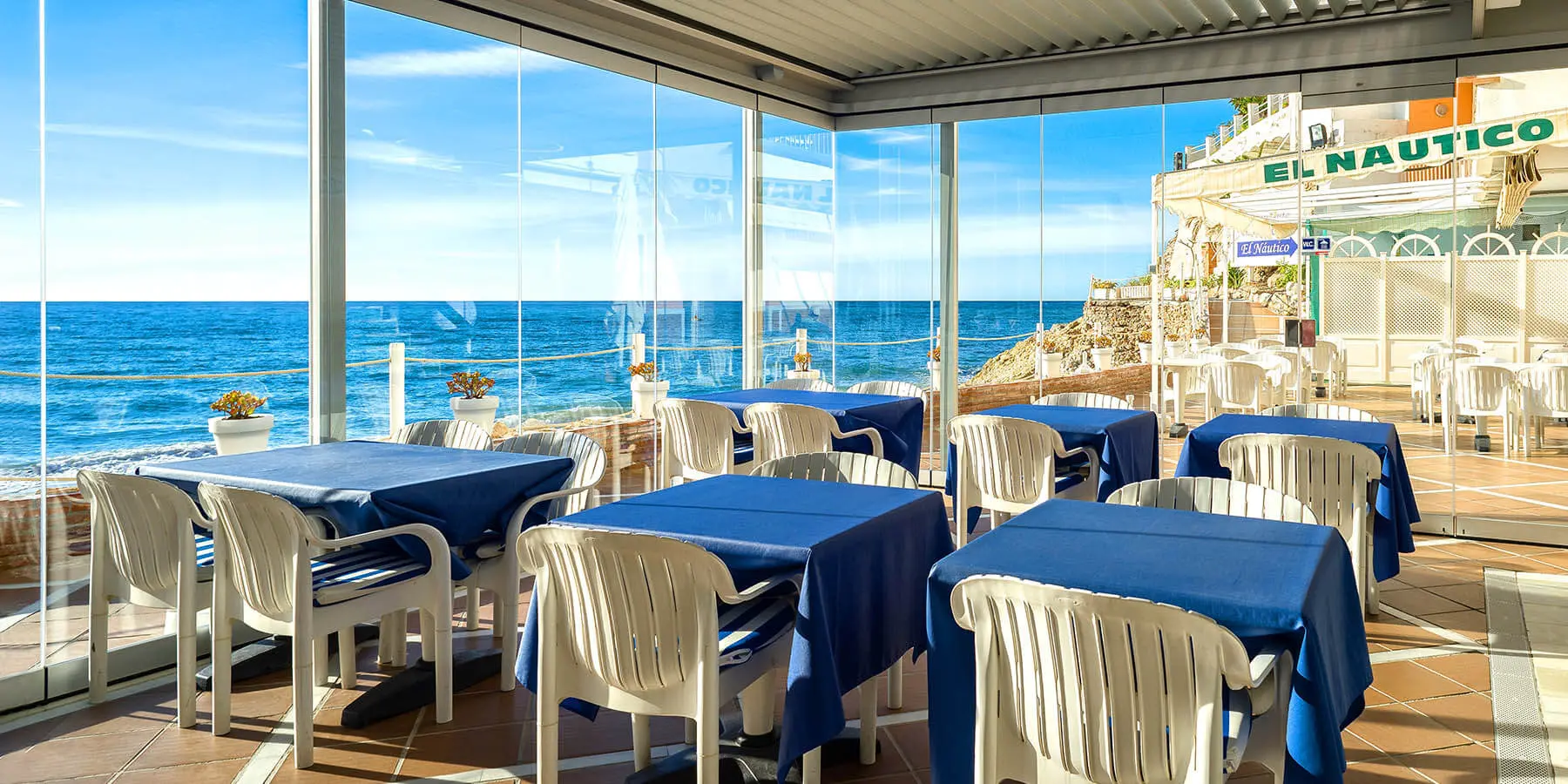 Restaurante con vista al mar, mesas con manteles azules y sillas blancas.