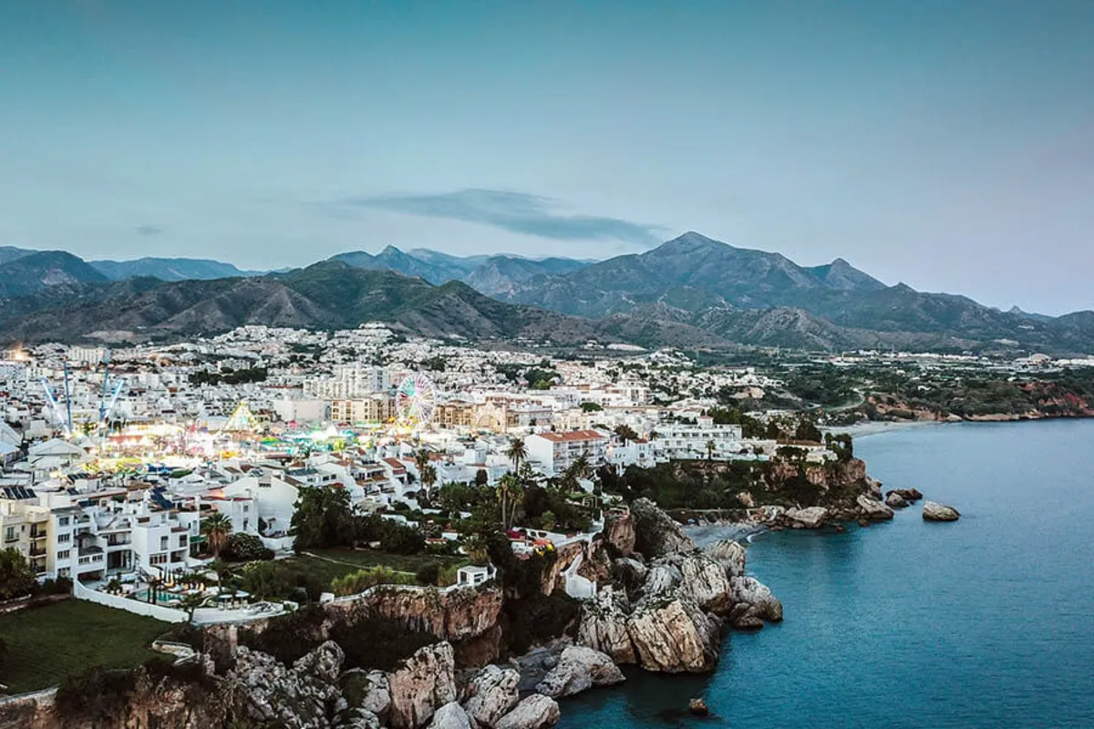 Vista a&eacute;rea de un pueblo costero con monta&ntilde;as al fondo y un mar azul.