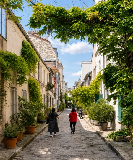 All&eacute;e pav&eacute;e entre maisons, arbres verts et deux personnes marchant vers le fond.