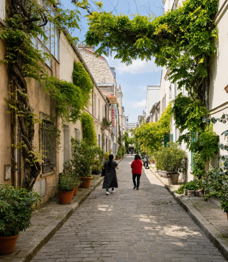 Ruelle pav&eacute;e bord&eacute;e d&rsquo;immeubles et de plantes vertes, deux personnes marchant