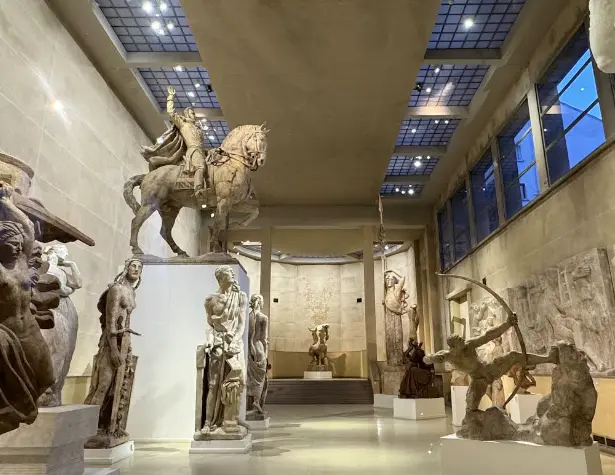 Galerie de mus&eacute;e avec sculptures antiques sur pi&eacute;destaux et vitrines.