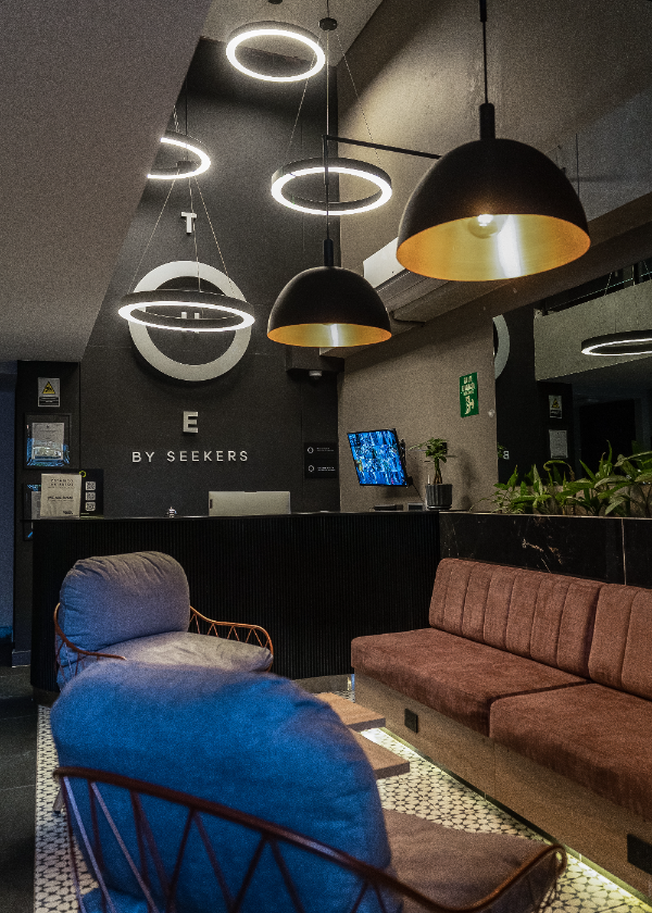 Lobby moderno con sillones grises y marrones, iluminaci&oacute;n circular y plantas.