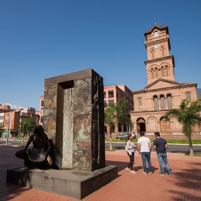 Edificio hist&oacute;rico y escultura en plaza peatonal con personas conversando.