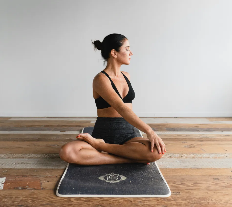 Mujer haciendo yoga sentada en un tapete sobre piso de madera.
