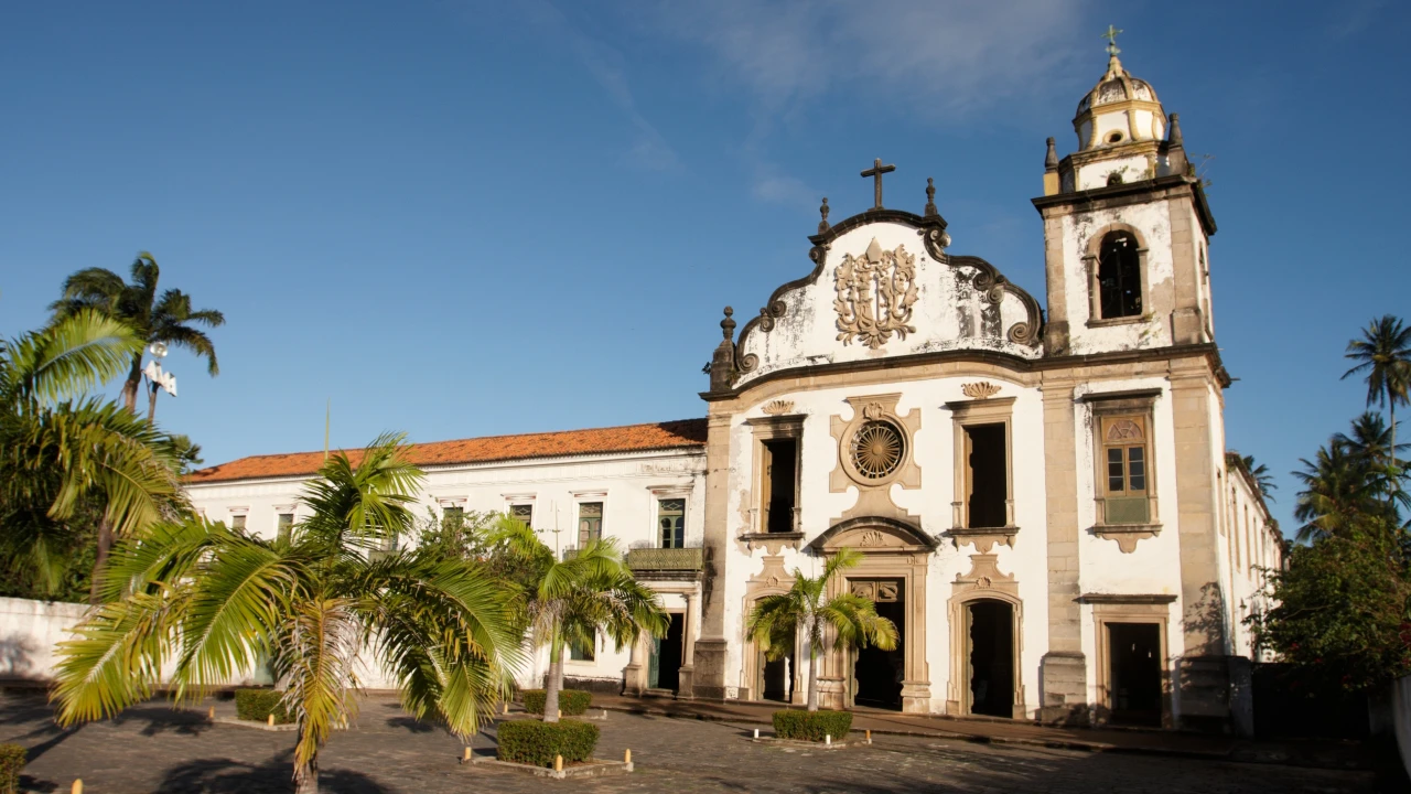 Igreja colonial com palmeiras em primeiro plano e c&eacute;u azul ao fundo.