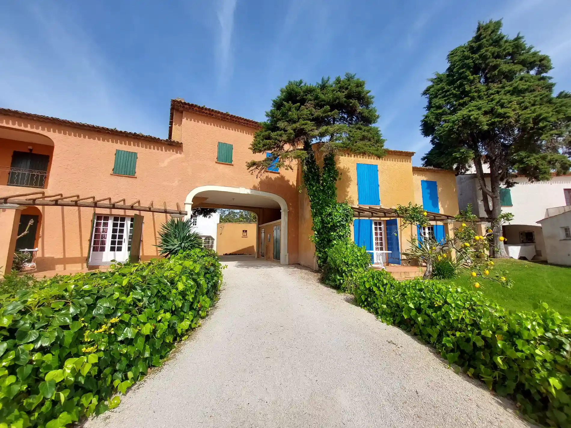 Casa laranja com janelas azuis, jardim e &aacute;rvores ao redor, sob c&eacute;u azul.