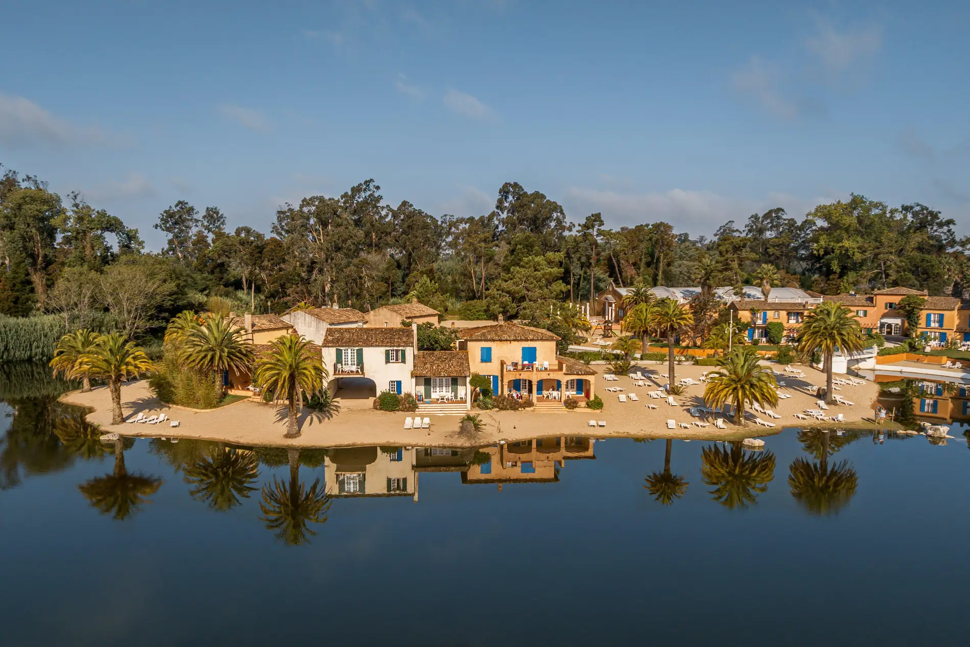 Casas em uma praia com palmeiras, refletidas na &aacute;gua calma.