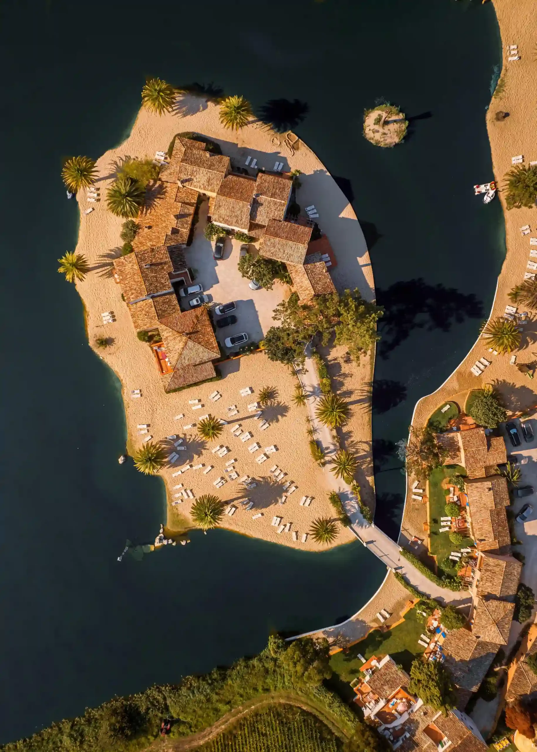 Vista a&eacute;rea de ilha com constru&ccedil;&otilde;es, praia e palmeiras, cercada por &aacute;gua.