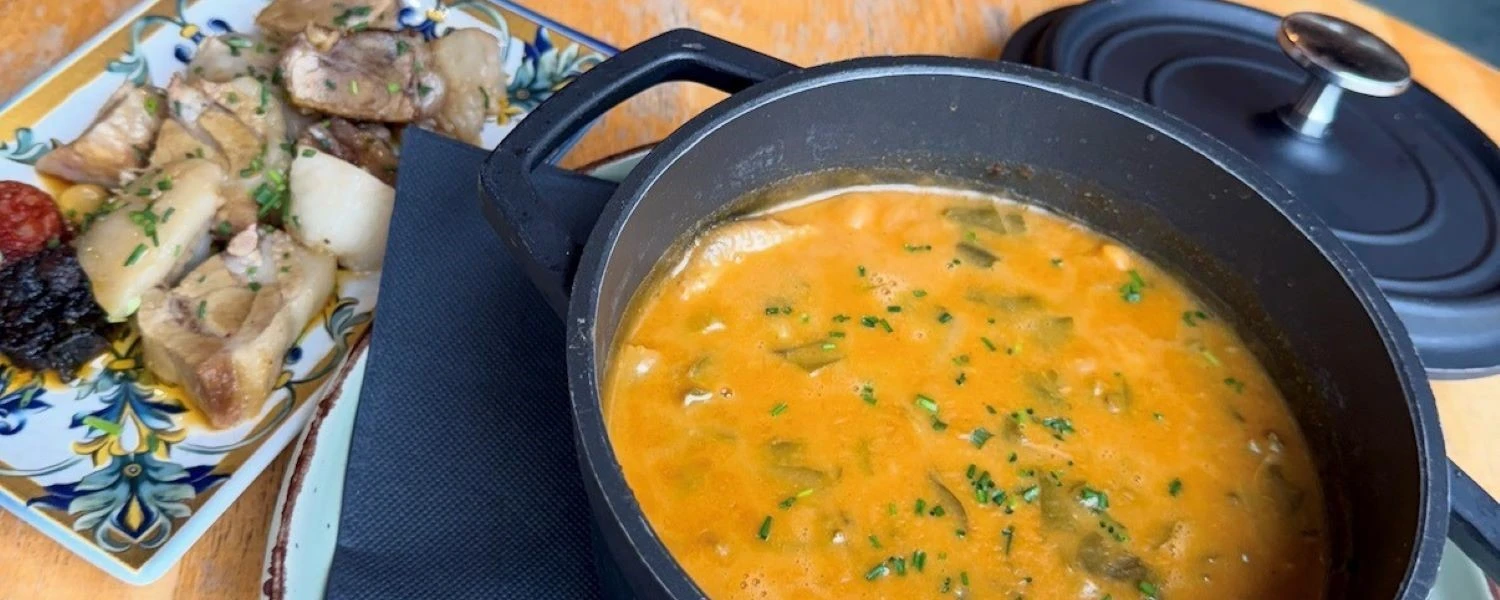 Sopa cremosa anaranjada en olla negra, con hierbas y plato con carne al lado