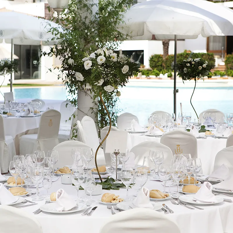 Mesa de banquete con cristaler&iacute;a y flores blancas bajo carpas junto a piscina