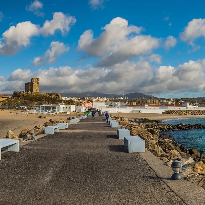 Camino costero con rocas, gente y un castillo junto al mar bajo nubes.