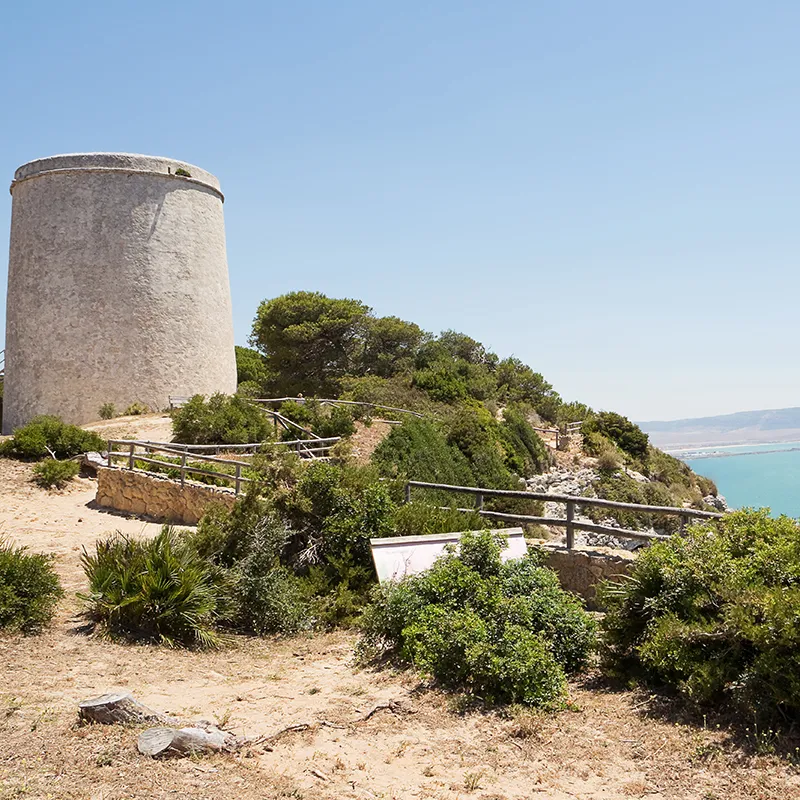 Torre cil&iacute;ndrica de piedra junto a un sendero con vegetaci&oacute;n costera
