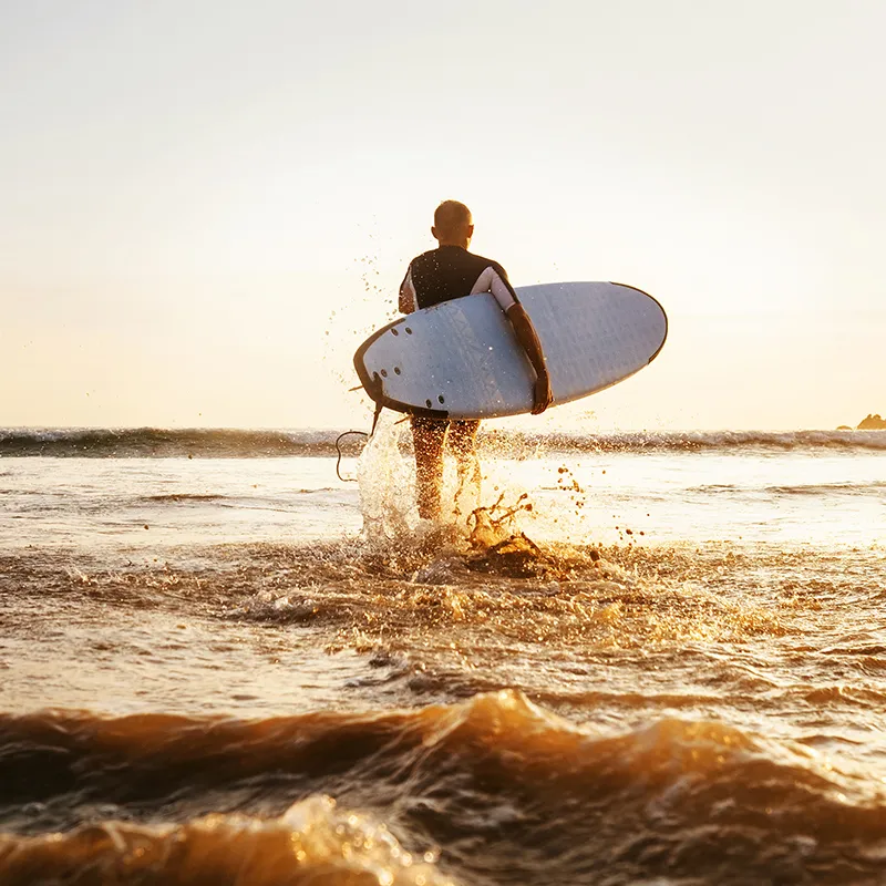 Surfer con tabla blanca saliendo del agua al atardecer, salpicaduras.