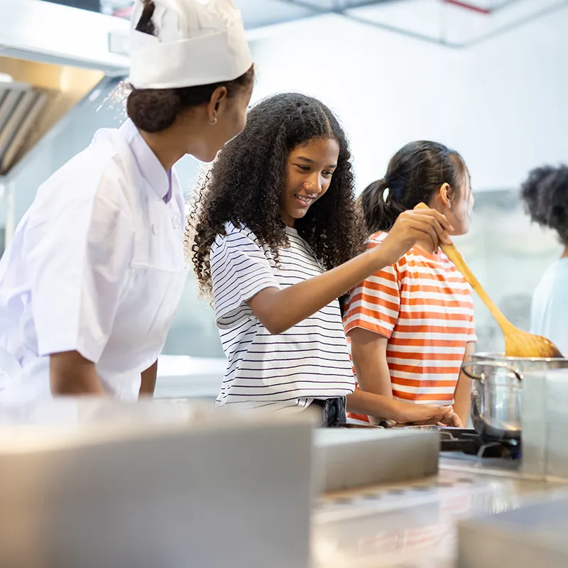 En cocina, una chef ayuda a un ni&ntilde;o a cocinar con una esp&aacute;tula.