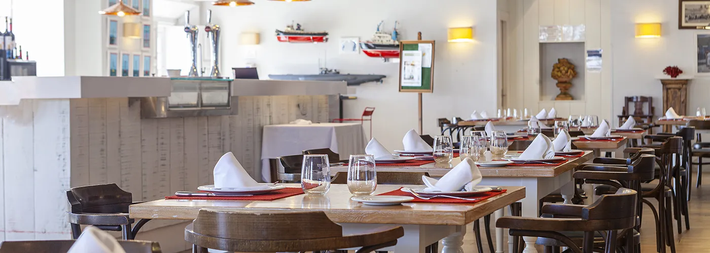 Comedor de restaurante con mesas puestas y cubiertos, luz c&aacute;lida y ventanas abiertas