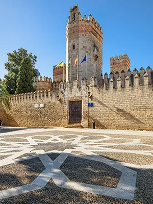 Torre de piedra y muralla medieval con calles y sombras bajo el sol.