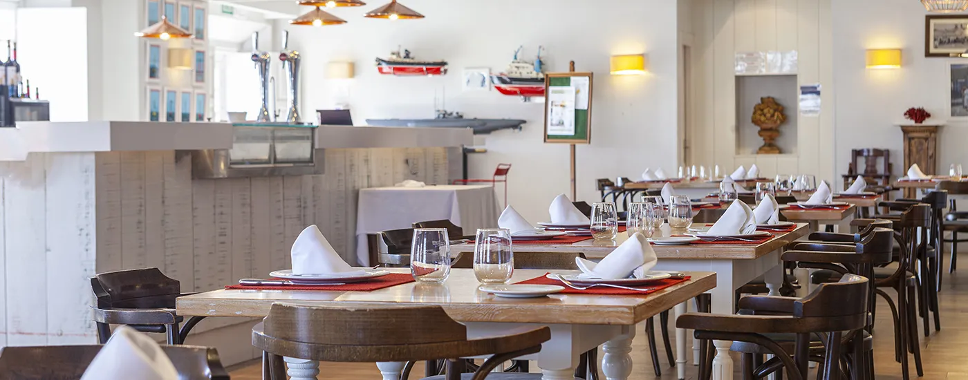Comedor de restaurante con mesas servidas y sillas, luces colgantes y vajilla.