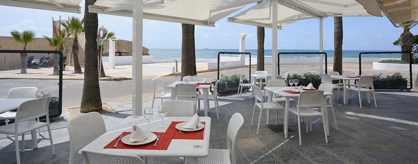 Terraza con mesas y sillas blancas bajo toldos, frente al mar.