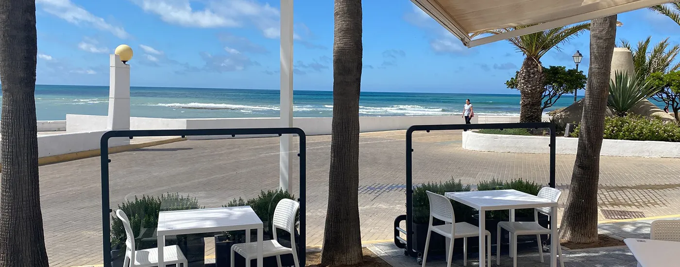 Terraza frente al mar con palmeras y mesa blanca bajo un toldo.