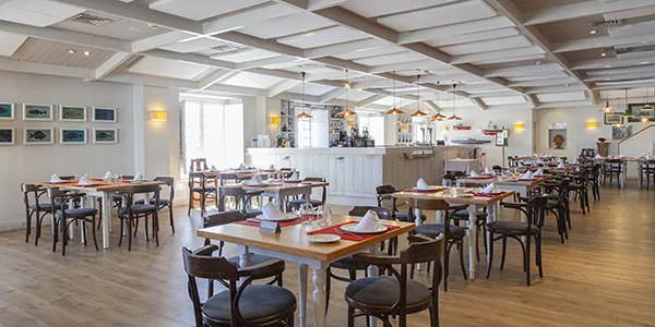 Comedor de hotel con mesas y sillas, iluminaci&oacute;n c&aacute;lida y buffet al fondo.
