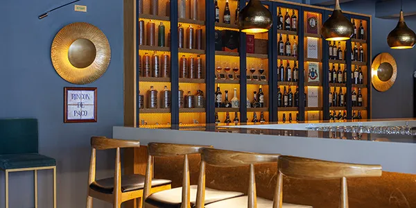 Bar con botellas en repisa dorada y sillas de madera en ambiente c&aacute;lido