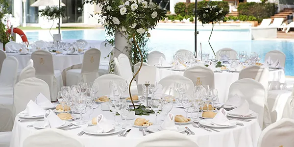 Mesa de banquete con manteles blancos, copas y flores junto a la piscina.