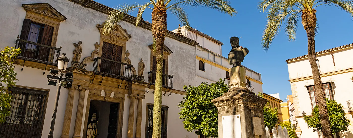 Estatua de bronce y palmeras frente a edificios hist&oacute;ricos con balcones, cielo azul